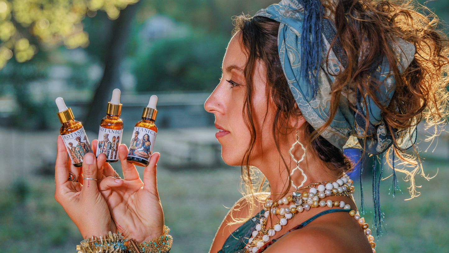 A woman in a blue bohemian-style dress and headscarf holds three small dropper bottles with amber liquid and gold caps, photographed outdoors in warm natural light with blurred trees in the background.