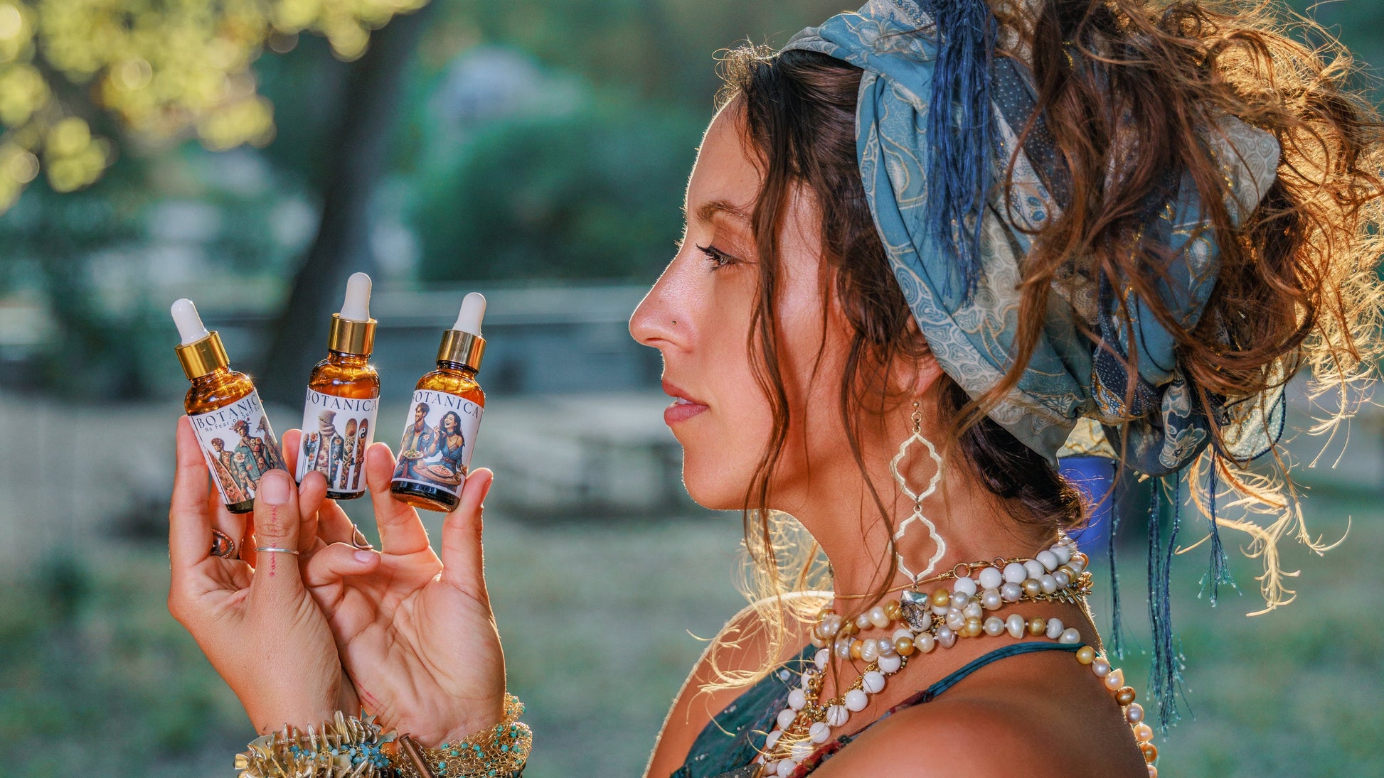 A woman in a blue bohemian-style dress and headscarf holds three small dropper bottles with amber liquid and gold caps, photographed outdoors in warm natural light with blurred trees in the background.